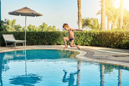 Young boy in swimming trunks jumping into childrens pool against sunbeds and palm trees in luxury hotel on warm summer evening in Greeceの写真素材