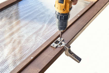 Installing of furniture hinge on wooden cabinet facade with glass using contemporary electric...