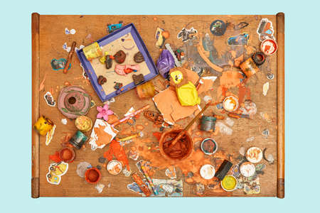 Jars of colorful paints brushes plasticine pieces and small toys on messy wooden table in light children room upper view isolated on blue backgroundの写真素材