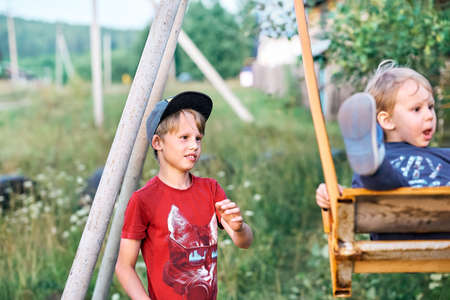 Smiling boy in cap pushes emotional junior brother on swings in green meadow near cottage in village on nice summer dayの写真素材