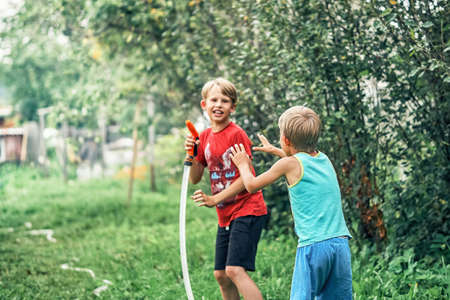 Joyful funny boy with cute little brother plays with water hose in green garden in spacious summerhouse yard on nice sunny dayの写真素材