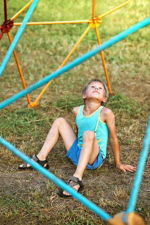 Adorable little boy in blue clothes sits on green grass under colorful climbing play set looking upward in city park on nice spring dayの写真素材