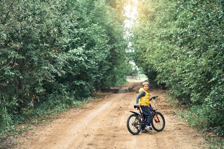 Adorable little boy in yellow vest with modern bicycle looks at camera on rural ground road with green trees on sides in summerの写真素材