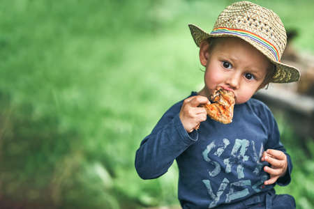 Adorable toddler boy in straw hat eats grilled chicken wing in green country house yard on summer day close view space for designの写真素材