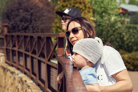 Cheerful young mother with cute children look at animals standing by high metal railing in large modern zoo on sunny dayの写真素材