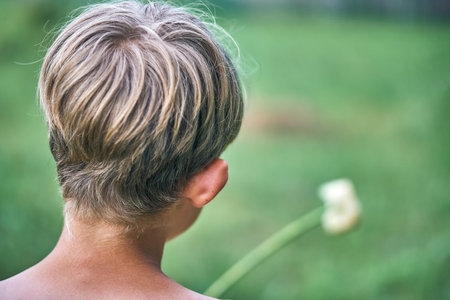 Blond boy roasts sweet marshmallow on stick above grill with cooking meat on green garden lawn on...