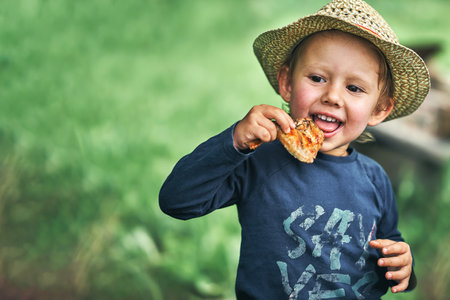 Adorable toddler boy in straw hat eats grilled chicken wing in green country house yard on summer day close view space for designの写真素材
