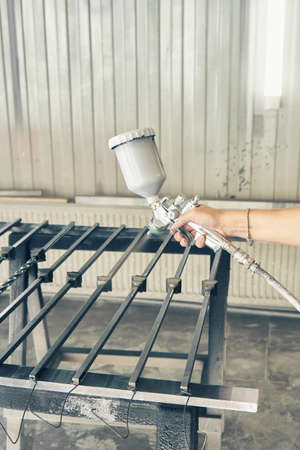 Woman worker paints forged balusters in black color with spray gun on rack support in contemporary spraying booth close viewの写真素材