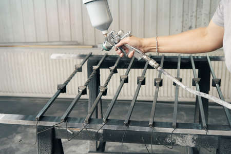Woman worker paints forged balusters in black color with spray gun on rack support in contemporary...