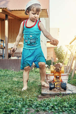 Barefoot little boy puts wet sand into bucket of yellow toy bulldozer on wooden board in country...