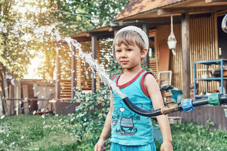 Blond little kid boy turns lever on tap with rubber hose and running up water playing in country house backyard on summer eveningの写真素材