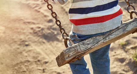 Little child in striped sweater and jeans plays old wooden swings on sand playground at sunset light close backside viewの写真素材