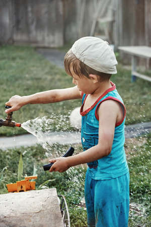 Blond little kid boy in a wet T-shirt turns lever on tap with rubber hose and running up water playing in country house backyardの写真素材