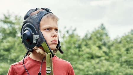 Frustrated teenage boy wearing vintage leather pilot helmet looks up into sky standing in green park on spring day close viewの写真素材