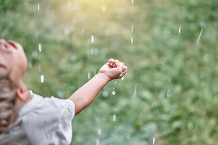 Cute little child boy enjoys warm summer rain catching falling drops with palms on green meadow in cottage backyard closeupの写真素材