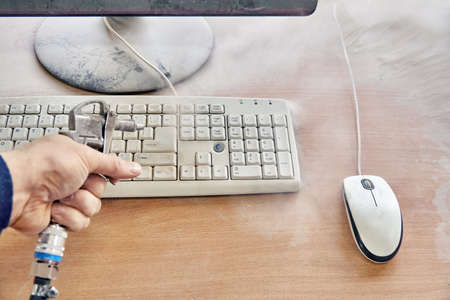 Employee cleans a dusty keyboard and dusty desk with air duster cleaning spray gun in the workshop...