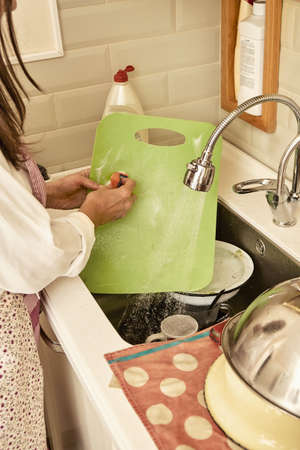 Woman in apron and foamy hands washes dirty green plastic cutting board with running water in modern...