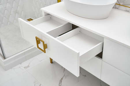 Elegant white cabinet with open drawer and shiny golden handles and leg with vessel sink in contemporary bathroom closeupの写真素材