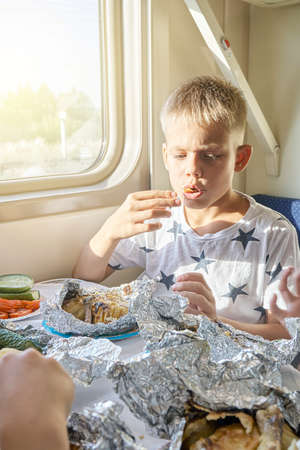 Blond teen boy have lunch eating fried chicken sitting at table in train car against landscape outside window closeupの写真素材