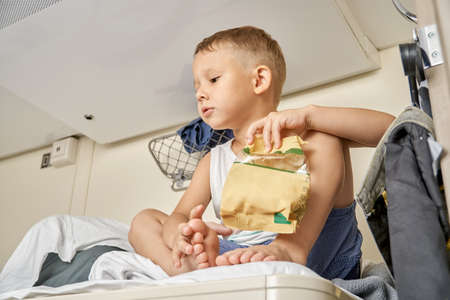 Blond little boy wearing vest and shorts eats snacks from paper pack resting on top shelf of train car traveling with family closeupの写真素材