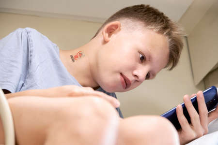 Teen boy with short blond hair reads electronic book with smartphone sitting on upper rack traveling in train car closeupの写真素材