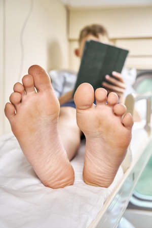 Bare feet of schoolboy lying on top rack bed with white bedding and playing games on tablet PC travelling by train extreme closeupの写真素材