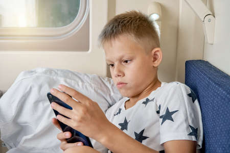 Teen boy with short blond hair reads electronic book with smartphone lying on white bedding on train car bottom shelf close shotの写真素材