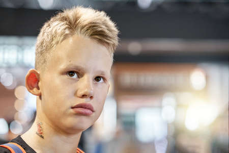 Bored blond-haired teenage boy with serious face expression sits in airport waiting hall against blurred crowd extreme closeupの写真素材