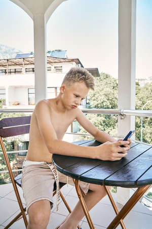 Blond-haired teenager boy wearing summer shorts plays online game sitting at wooden table on hotel balcony at bright sunlight closeupの写真素材