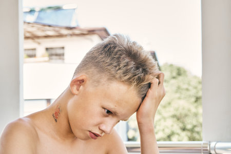 Teenager boy with short blond hair and bare shoulders props head with hand sitting on balcony of hotel against green trees closeupの写真素材