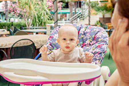 Small boy sits in baby armchair of outdoor restaurant under mother care. Child plays with blue pen against colorful and blooming garden plantsの写真素材