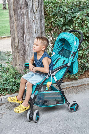 Preschooler frowns sitting in blue stroller of baby brother walking in park. Fair-haired boy sits and rests tired from long stroll on summer holidayの写真素材