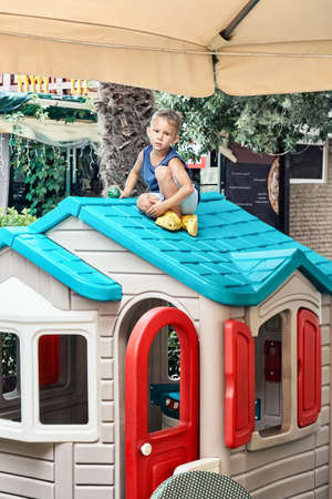 Preschooler boy in summer clothes sits on bright roof of toy house in restaurant play area. Fair-haired child spends free time playing outdoorsの写真素材