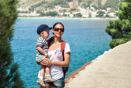 Young mother holds in arms little son in cap standing near trees against blue sea in coastal town Omis. Woman in sunglasses and toddler rest in Croatiaの写真素材