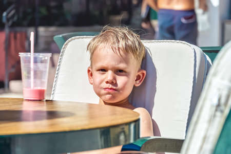 Cute blond boy sits in chair at table drinking milkshake in street cafe near beach in Omis. Toddler enjoys vacation in Croatia on sunny day closeupの写真素材