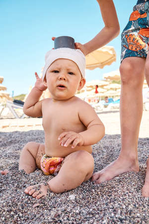 Elder brother plays with baby boy sibling sitting on sand in shadow from umbrella. Blond schooler and toddler in hat enjoy vacation on beachの写真素材