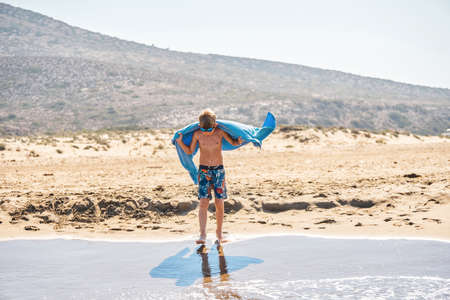 Junior schoolboy covered with blue towel walks on wet sand of wild beach. Blond boy in goggles stands on water edge looking at feet against high hillsの写真素材