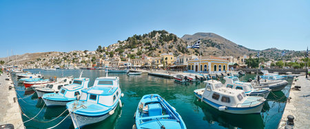 Symi, Greece - July 05 2017: Moored tourist and fishing boats at pier in port of island Symi in Greece. Vessels drift on turquoise water against small multicolored houses on hillsのeditorial素材