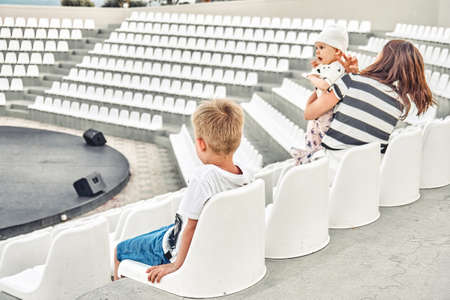Young mother with sons seats in empty amphitheate rwith white plastic seats for evening show at hotel. Schoolboy shows performance for familyの写真素材