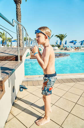 Schooler shivers with cold after swimming in pool with clear blue water on territory of hotel. Cute little blond boy with goggles on summer vacation at resortの写真素材