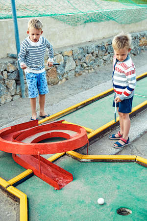 Blond brothers play children golf holding clubs at hotel playground. Boys playing golf watch ball to roll straight into hole after successful hitの写真素材
