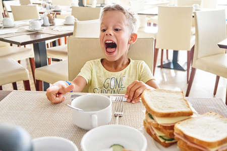 Blond preschooler boy laughs while having breakfast at hotel. Cute boy has fun sitting at table and eating big sandwiches on summer vacationの写真素材