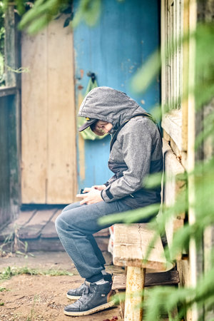 Focused schoolboy in warm jacket uses smartphone sitting on wooden bench near house in countryside....