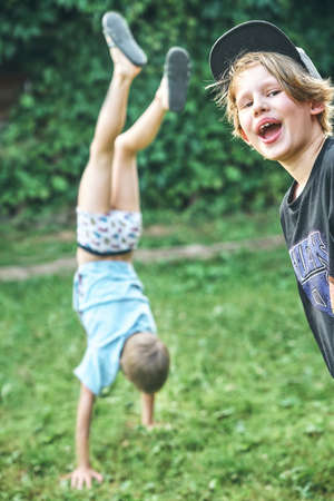 Cute boy in hat looks in camera against blurred view of elder brother making handstand on grass....