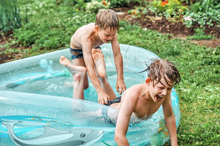 Brothers wrestle in children inflatable pool in cottage yard against lush greenery and flowers. Cheerful schoolboys have fun in water on summer in countryside. No logoの写真素材