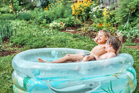Brothers wrestle in children inflatable pool in cottage yard against lush greenery and flowers. Cheerful schoolboys have fun in water on summer in countryside. No logoの写真素材