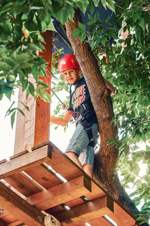 Boy in a helmet and climbing equipment standing on a wooden platform against foliage background, spending time in rope park in summer eveningの写真素材