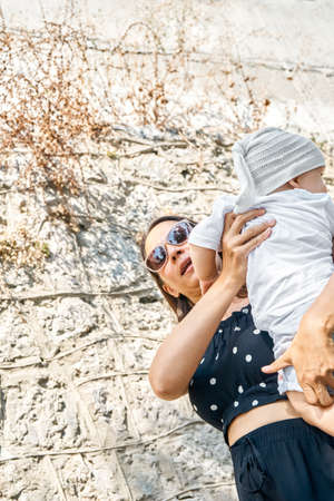 Mother in sunglasses holds cute toddler in arms against the wall. Young brunette woman and little boy enjoy summer vacation low angle close viewの写真素材