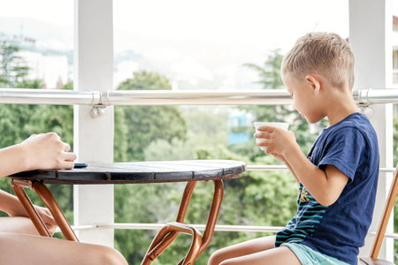 Preschooler boy drinks warm milk from mug sitting on chair in early morning. Excited child enjoys summer holidays drinking beverage on hotel balconyの写真素材