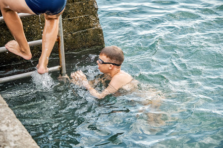 Schoolboy climbs up pier on metal ladder against seascape. Waves hit stone pier. Boy enjoys spending holidays swimming in sea closeupの写真素材